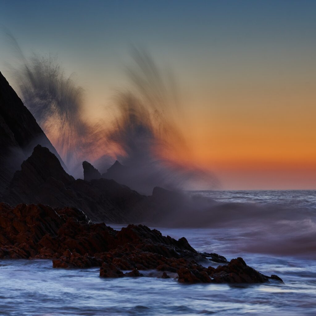 Ocean waves crashing on jagged rocks at sunset with vibrant sky and dynamic surf motion blur Wallpaper