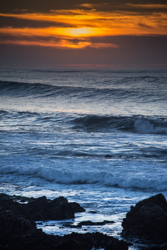 Ocean waves crashing on rocky shore at vibrant orange sunset coastal landscape wallpaper background
