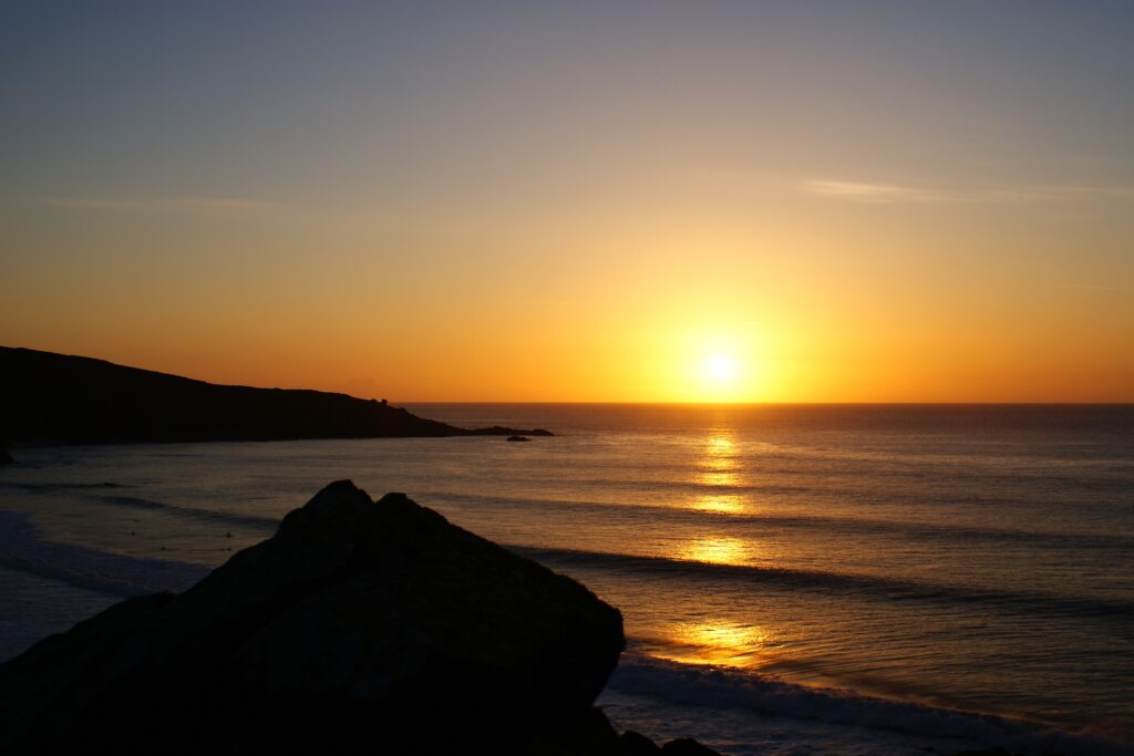 Sunset wallpaper with golden sun rays over calm ocean waves and silhouetted rock foreground