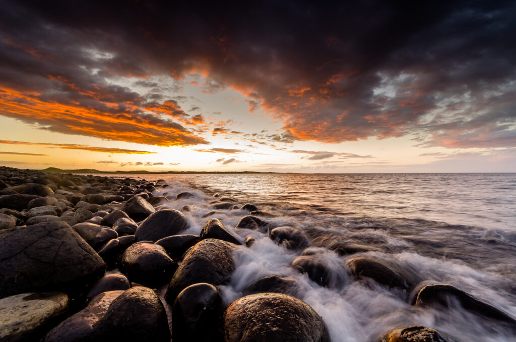 Sunset over ocean with orange gold clouds reflecting on waves and dark smooth coastal stones Wallpaper