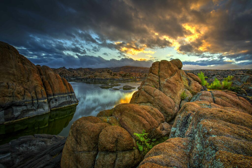 Sunset over rugged orange rocks and stone formations by a tranquil river with dramatic mountains Wallpaper