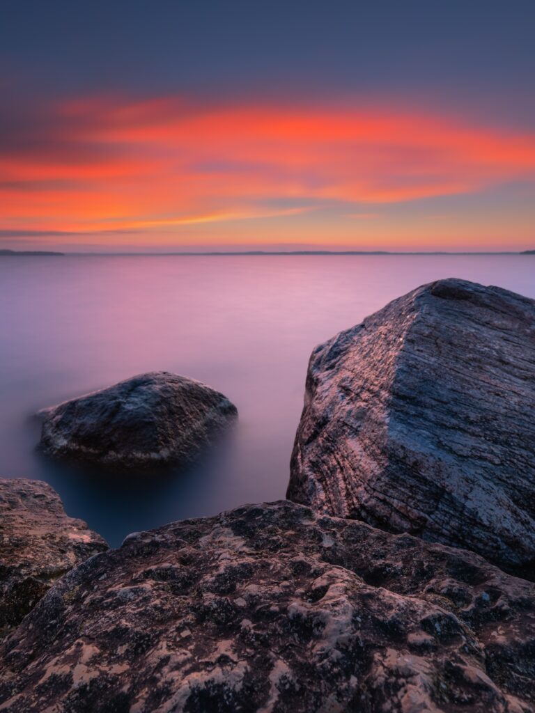 Rugged seaside rocks at sunset with vibrant orange and purple sky over calm water wallpaper