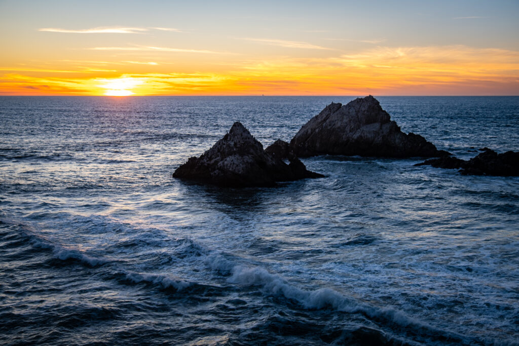 Sunset over rugged sea rocks with gentle waves blending warm golden and cool blue tones Wallpaper