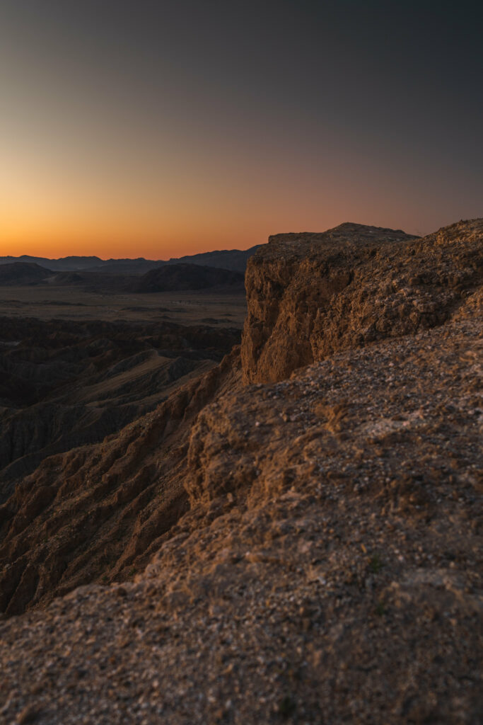 Sunset over rugged stone cliffs and rocky terrain with distant mountains under a clear sky Wallpaper