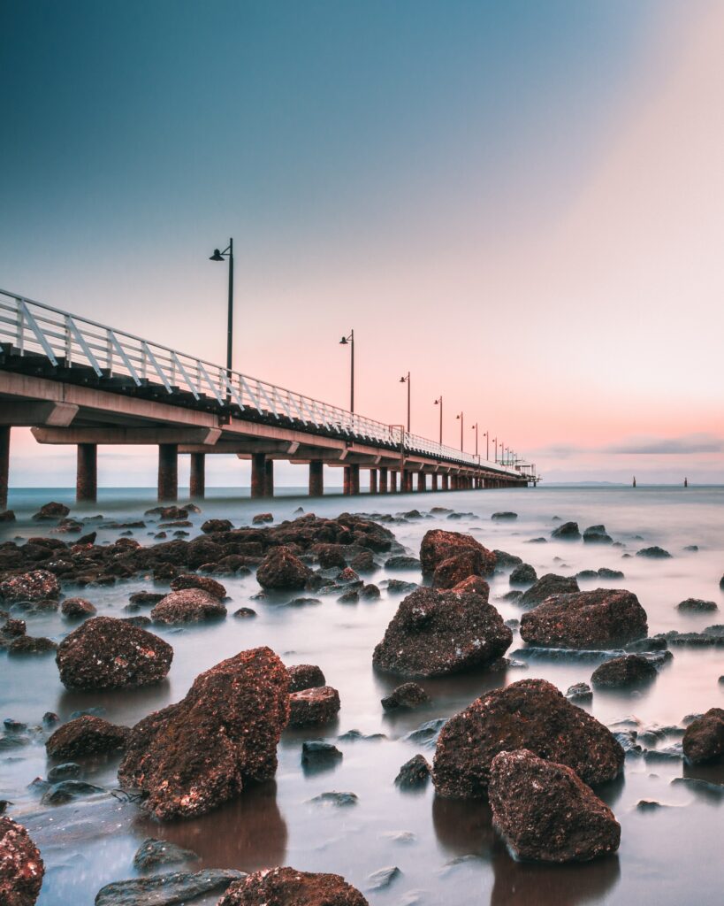Serene sunset pier over calm sea with pastel horizon and textured stones foreground wallpaper