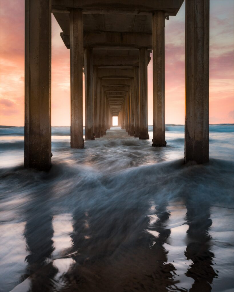 Sunset view beneath pier with sturdy pillars reflecting in swirling sea waves and pastel skies Wallpaper