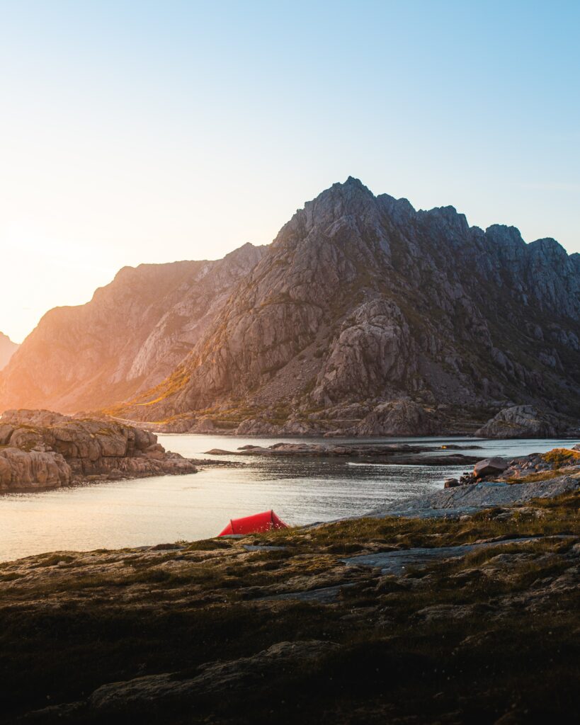 Vibrant red tent by tranquil lake with rugged rocks and mountains at sunset outdoor adventure Wallpaper