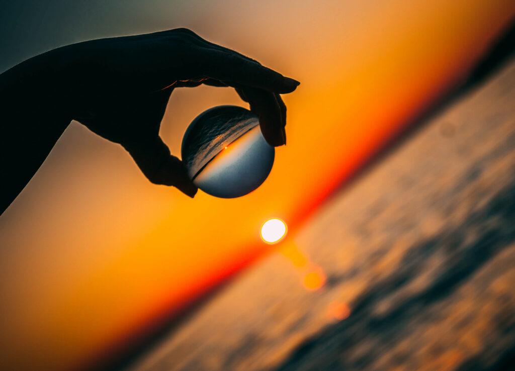 Silhouetted hand holding glass ball over calm ocean reflecting vibrant orange sunset sky Wallpaper