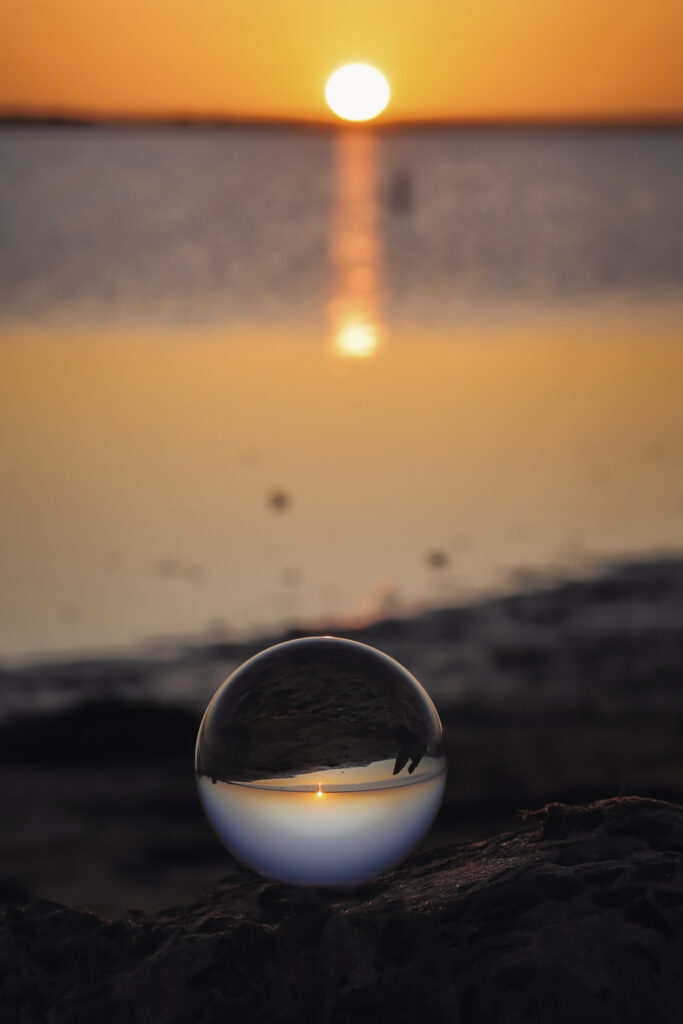Glass ball on sand reflecting golden sunset over calm water serene nature wallpaper background