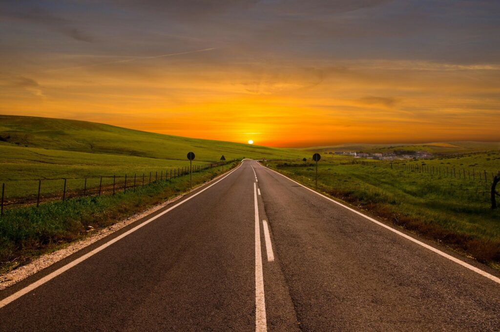 Asphalt road winding through green fields at sunset with warm orange and golden sky colors Wallpaper