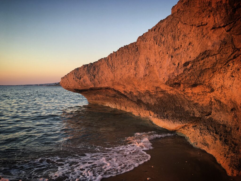 Sunset rocky coastline with textured cliffs and gentle waves blending earth tones and blue sea Wallpaper
