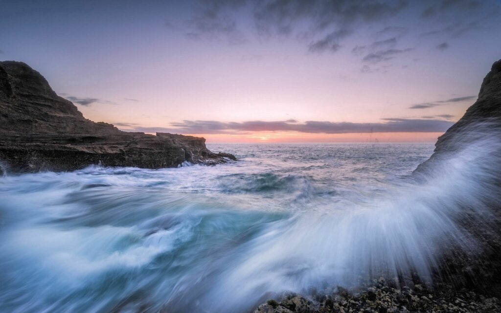 Sunset seascape with vibrant waves crashing on rugged rocks under a pastel blue and pink sky Wallpaper