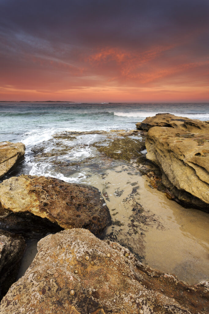 Dramatic sunset seascape with rugged shore rocks gentle waves and sandy beach wallpaper background