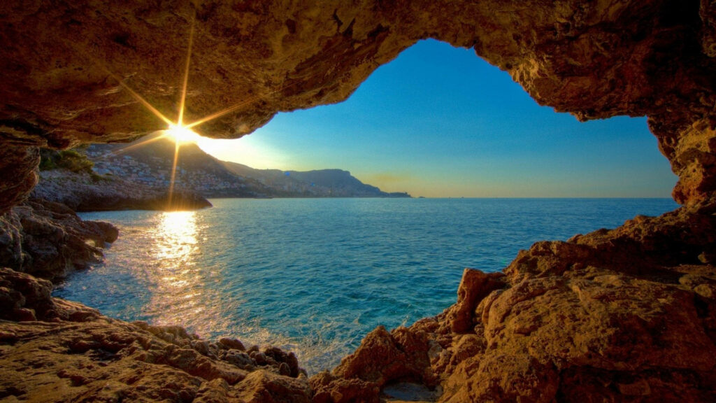 Golden sunset over Mediterranean sea framed by rugged cave with distant coastal town view Wallpaper