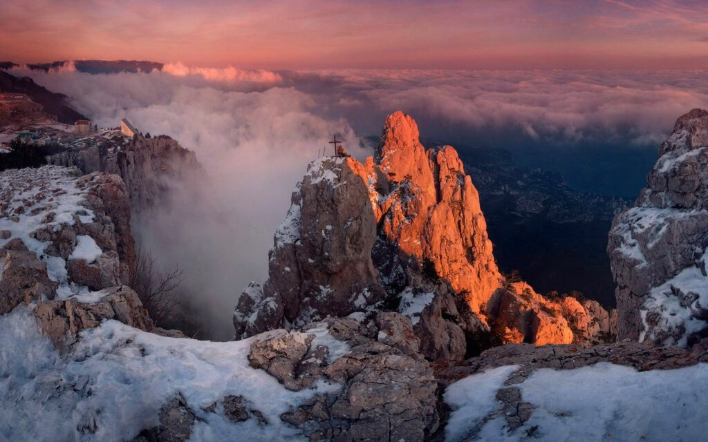 Snow-covered mountain peak with cross monument at sunset misty clouds and distant city valley Wallpaper