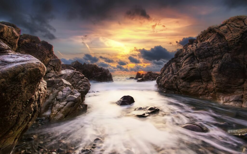 Sunset landscape with rugged rocks beside serene stream flowing to sea under dynamic clouds Wallpaper