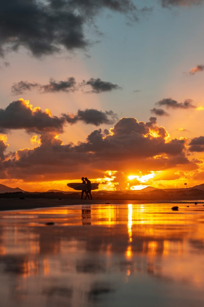 Silhouettes of surfers walking along ocean shore at sunset with warm orange gold sky reflection Wallpaper