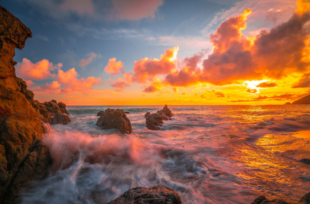 Golden sunset over rocky coastline with vibrant orange clouds and crashing ocean waves wallpaper