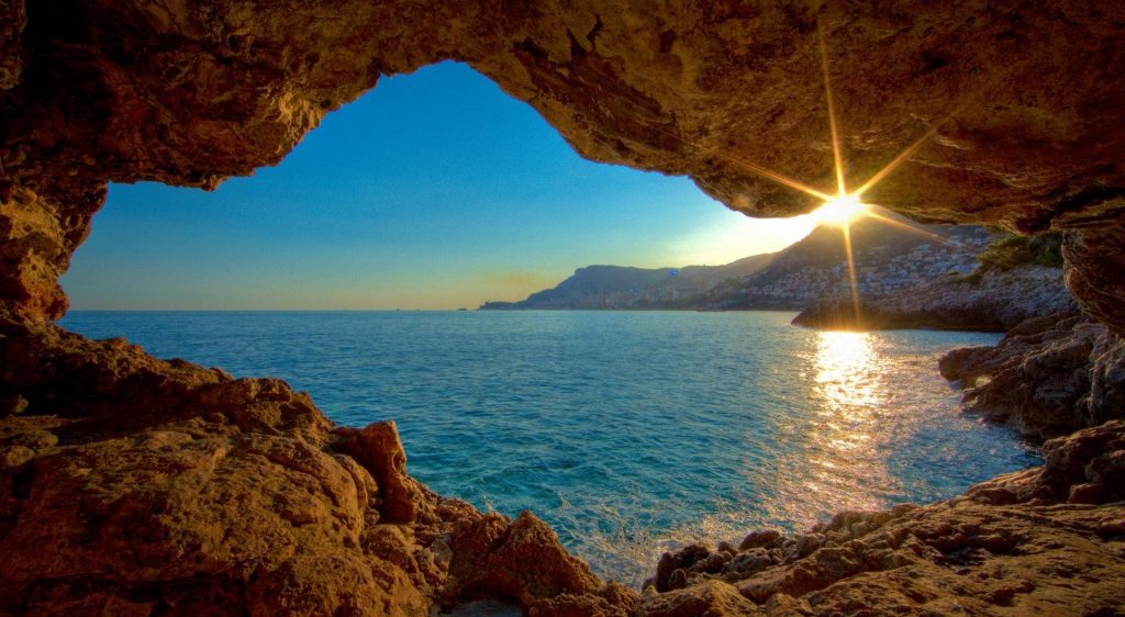 Sunshine wallpaper with rugged rock cave framing Mediterranean coastline and radiant sunburst