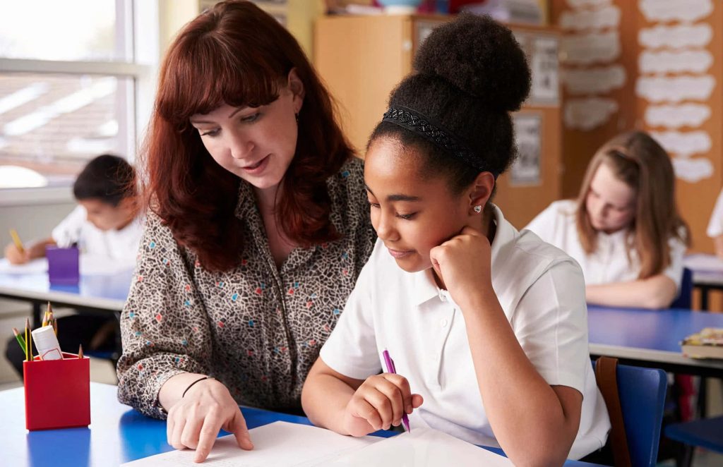 Teacher helping young student with notebook in bright classroom for academic mentorship wallpaper