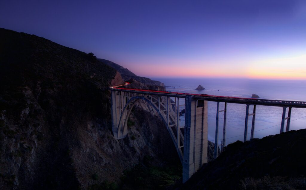 Sweeping coastal bridge with car light trails over cliffs and calm sea under deep blue night sky Wallpaper