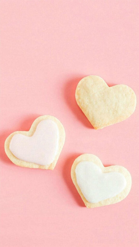 Three heart shaped cookies iced white and pink on pastel pink background minimal wallpaper