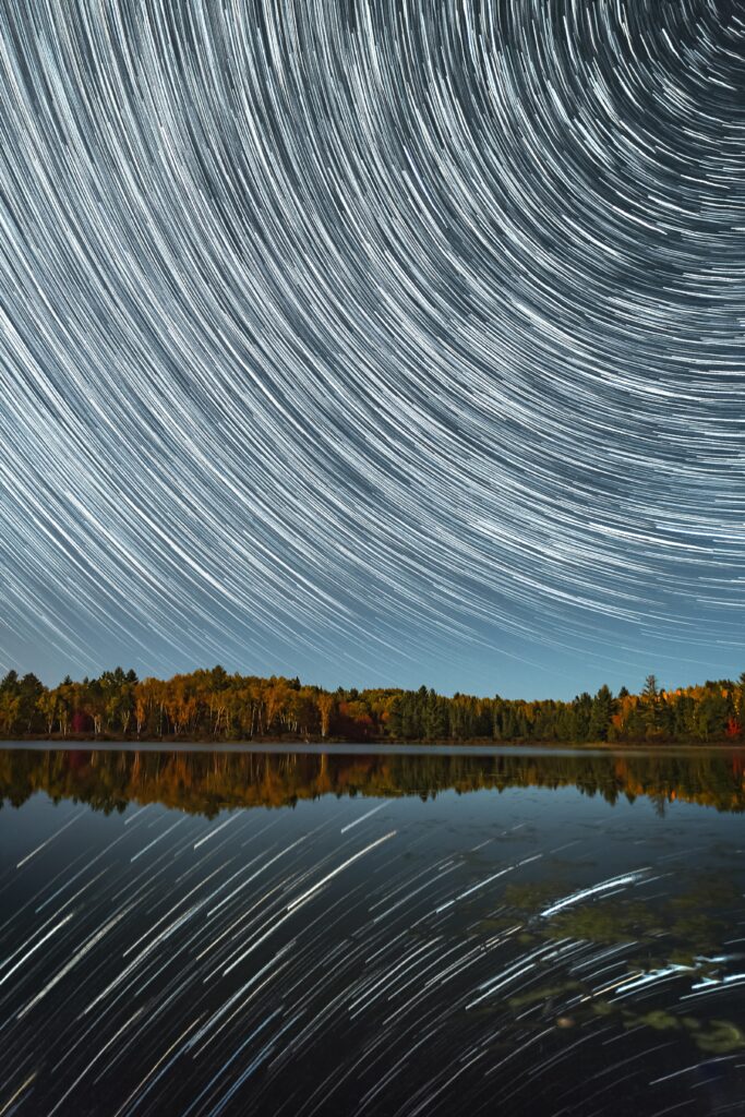 Long exposure star trails over autumn trees reflected on a calm lake under vibrant night sky Wallpaper