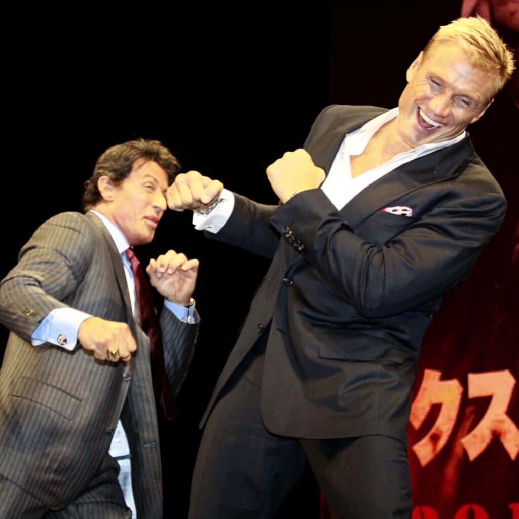 Sylvester Stallone and Dolph Lundgren in sharp suits share a dynamic playful moment wallpaper