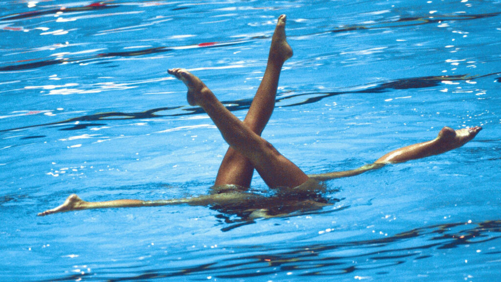 Two synchronized swimmers in artistic ballet leg pose above water in a clear blue pool Wallpaper