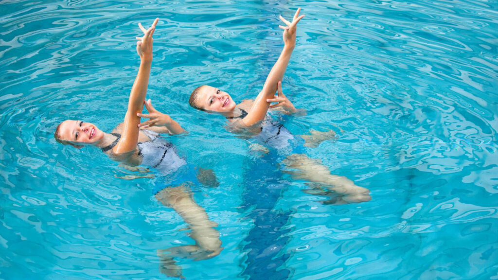 Two synchronized swimmers in elegant poses perform a routine in crystal-clear blue water background Wallpaper