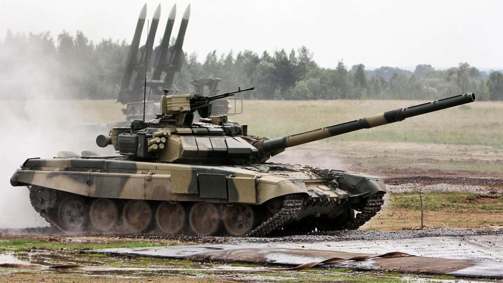 T-80U main battle tank charges on dusty field with Buk-M2 missile system in overcast light Wallpaper