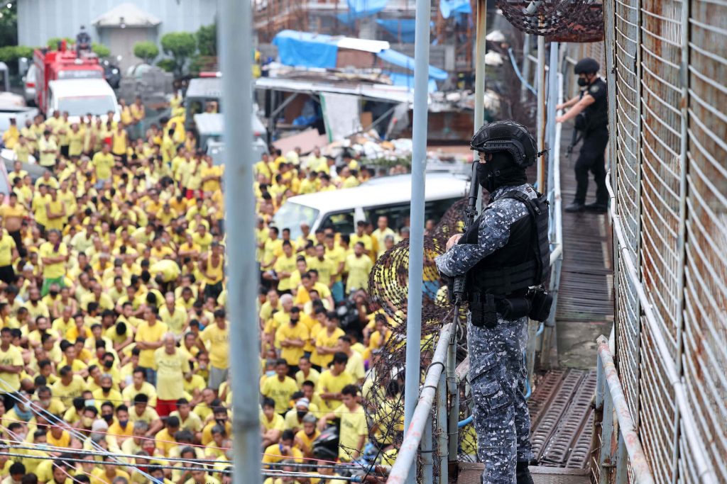 Tactical guard in camouflage watches crowded Philippine prison yard with inmates in yellow shirts Wallpaper