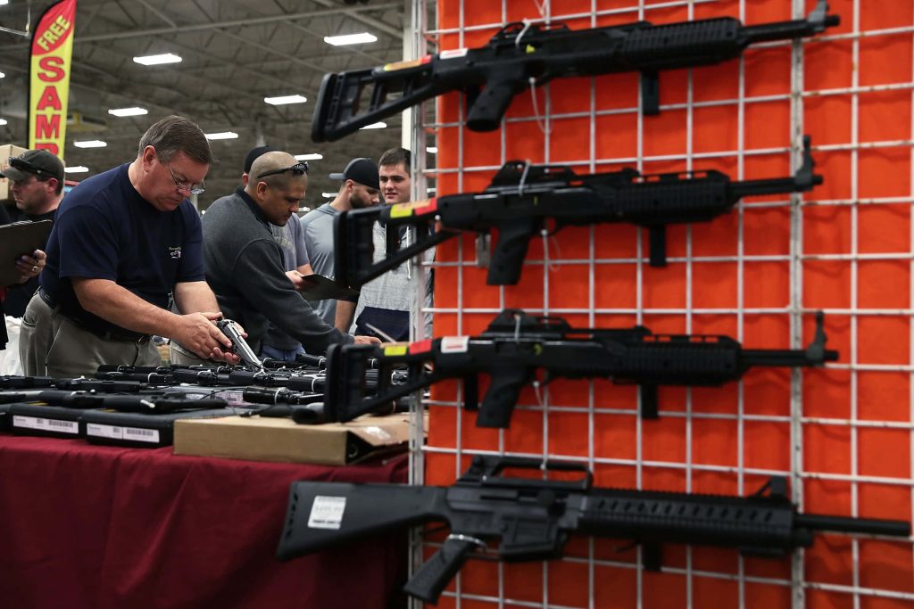 Busy gun show with tactical rifles on orange rack and handguns inspected on table indoors Wallpaper
