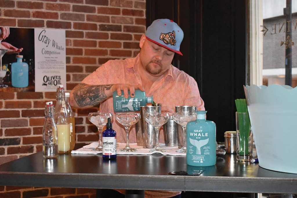 Tattooed bartender in blue cap pouring Gray Whale Gin in stylish bar with cocktail tools and event poster Wallpaper