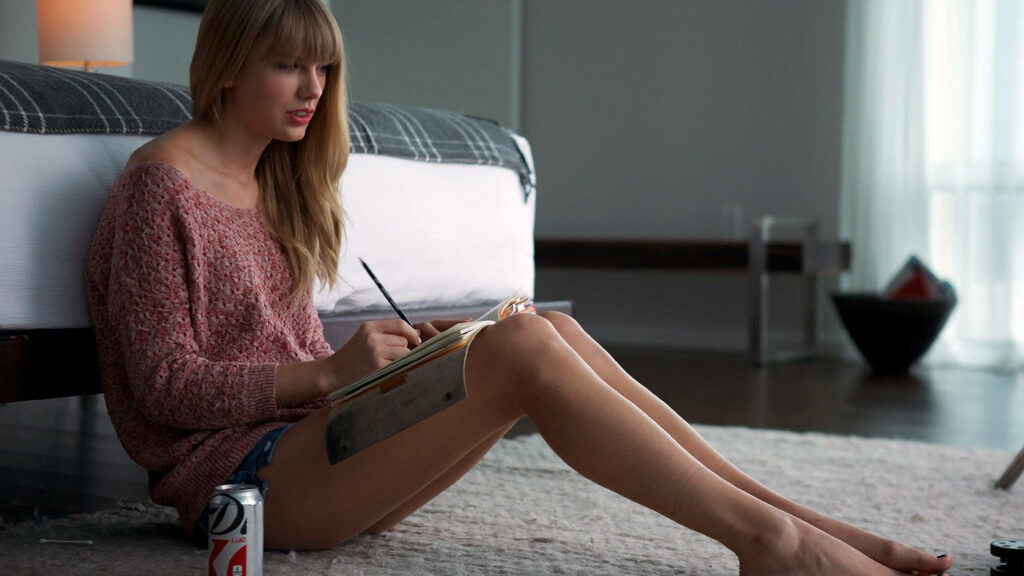 Taylor Swift sitting on floor writing in notebook wearing pink sweater and denim shorts cozy room Wallpaper