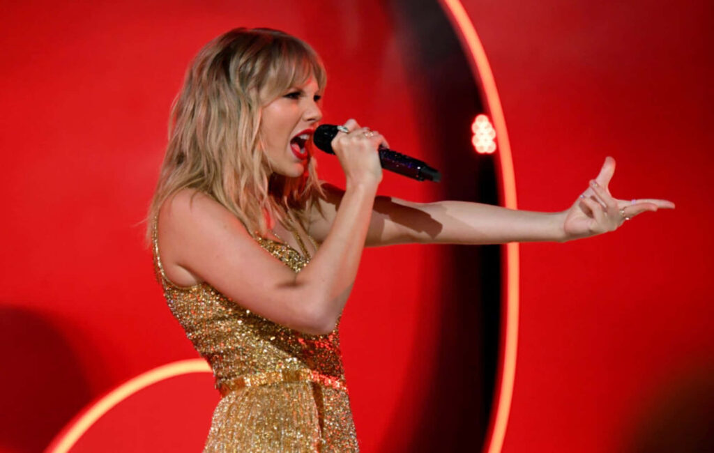 Taylor Swift performing live in sparkling gold dress vibrant red backdrop Red Taylors Version album Wallpaper