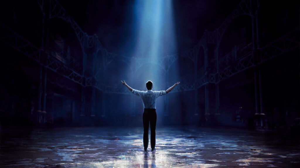 Man center stage in spotlight in grand dark hall showcasing The Greatest Showman live show energy Wallpaper