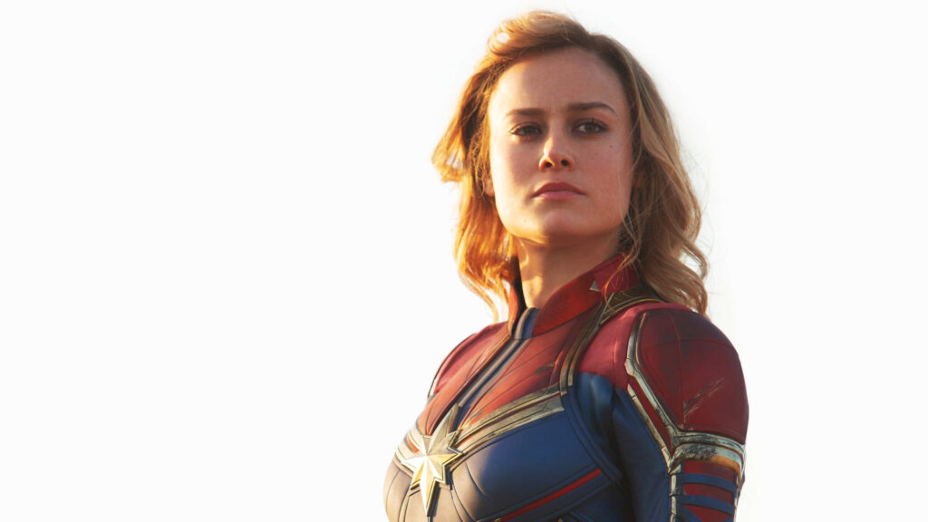 The Mighty Heroine: Brie Larson as the Unstoppable Captain Marvel, Radiant in Her Iconic Red, Blue, and Gold Attire, Set Against a Mesmerizing Background Wallpaper