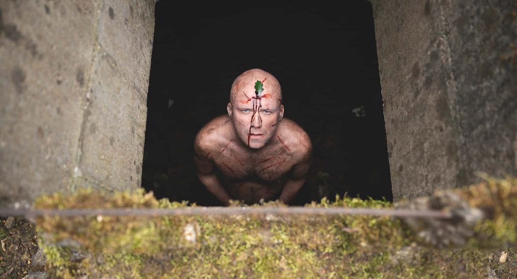 Shirtless bloodied man with leaf on forehead emerging from dark mossy bunker horror wallpaper