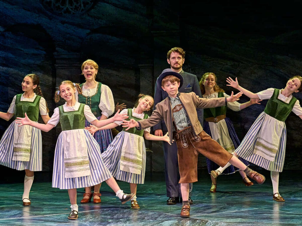 The Sound Of Music stage cast in traditional costumes singing and dancing under bright lights Wallpaper
