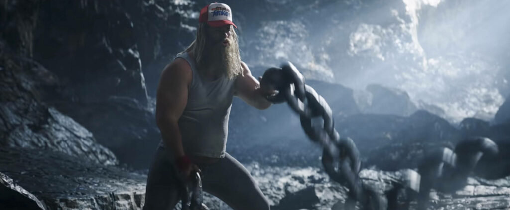 Thor in rocky cave wearing tank top and trucker hat wielding heavy chains from Love and Thunder Wallpaper