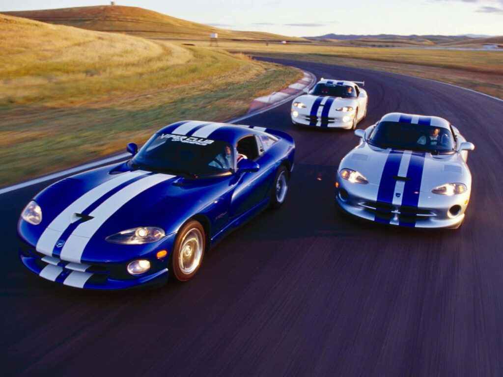 Three Dodge Vipers with blue and white stripes race on winding track by grassy hills at dusk Wallpaper