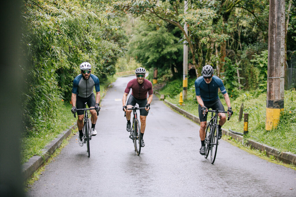 Three men in cycling gear ride racing bikes on a wet green countryside road under trees Wallpaper
