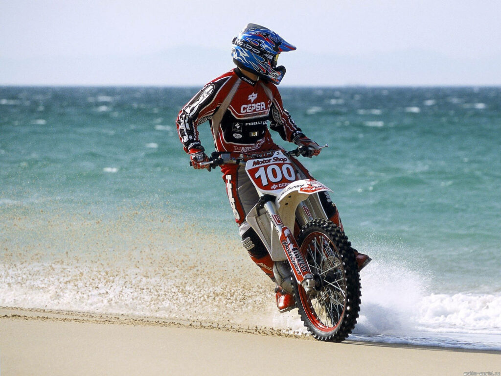 Dirt bike rider in red and blue gear speeds on sandy beach with turquoise ocean waves background Wallpaper