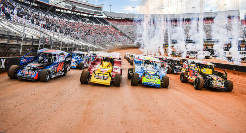 Vibrant stock cars racing on red clay track with dust and fireworks in packed stadium stands Wallpaper