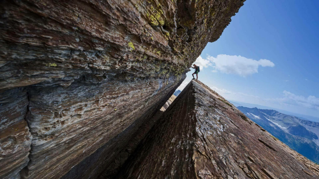 Climber ascending steep triangular mountain slope with rugged rocks and bright blue sky background Wallpaper