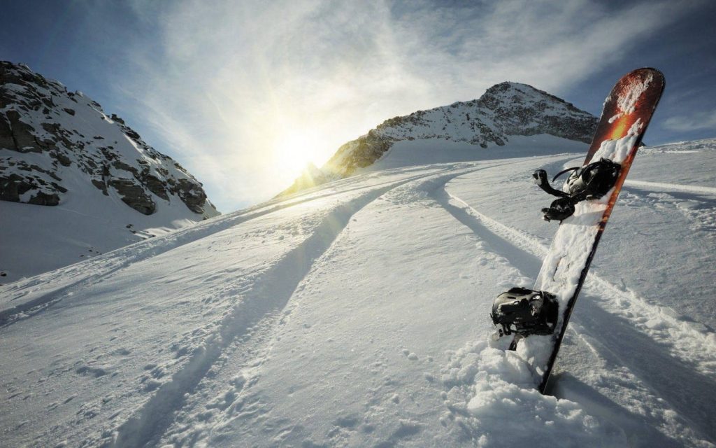 Snowboard upright in fresh snow with sunlit mountain peaks and ski tracks on pristine slope Wallpaper
