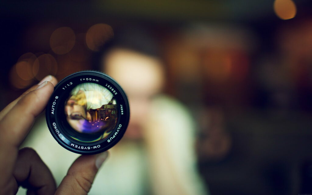Hand holding vintage Olympus OM-System 55mm f1.2 lens with sharp inverted reflection and bokeh Wallpaper