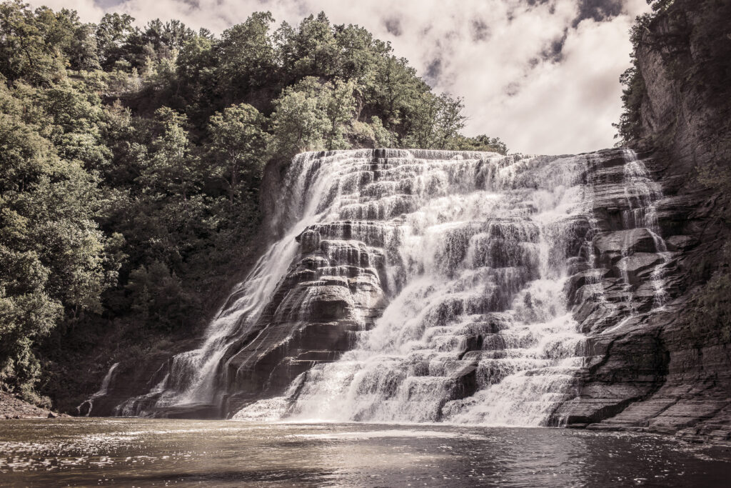 Powerful waterfall over layered rocks flows into river with lush trees steep cliffs sepia tone wallpaper