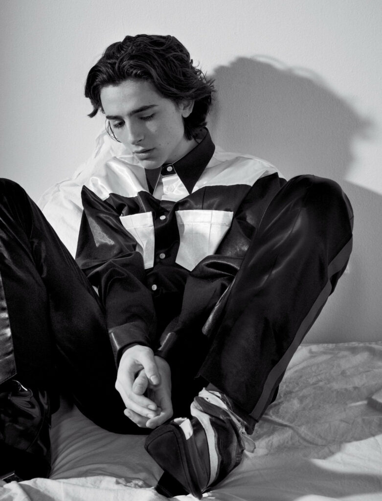 Timothée Chalamet stylish outfit greyscale photo sitting on bed contemplative soft lighting wallpaper